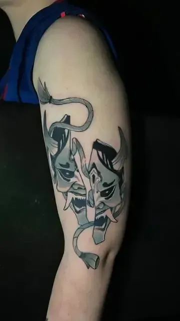 Tatuaje de dos máscaras de demonio japonesas, gris y blanca, en la parte superior del brazo de una persona