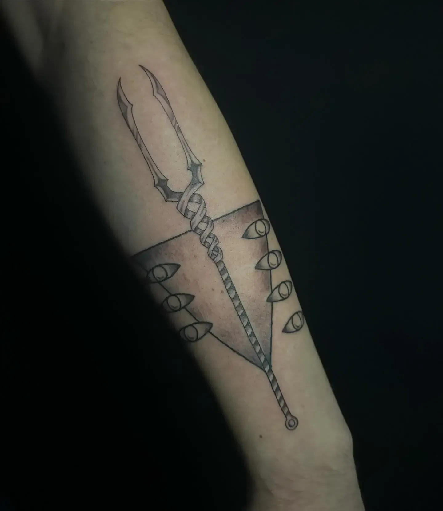 Tatuaje de la Lanza de Longino y múltiples ojos en un brazo, con fondo oscuro.