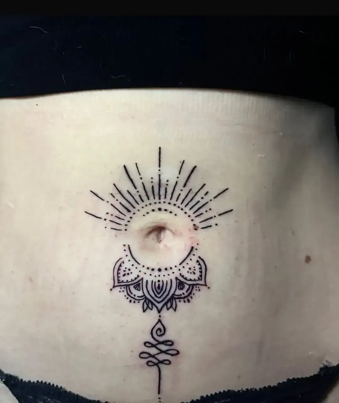 Tatuaje en el abdomen, con un diseño de rayos de sol sobre una flor de loto y un símbolo estilizado debajo.