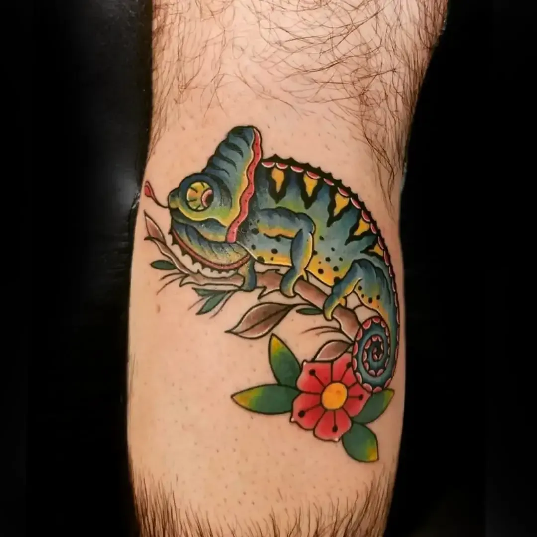 Tatuaje de un camaleón colorido en una rama con una flor roja, en un brazo peludo.