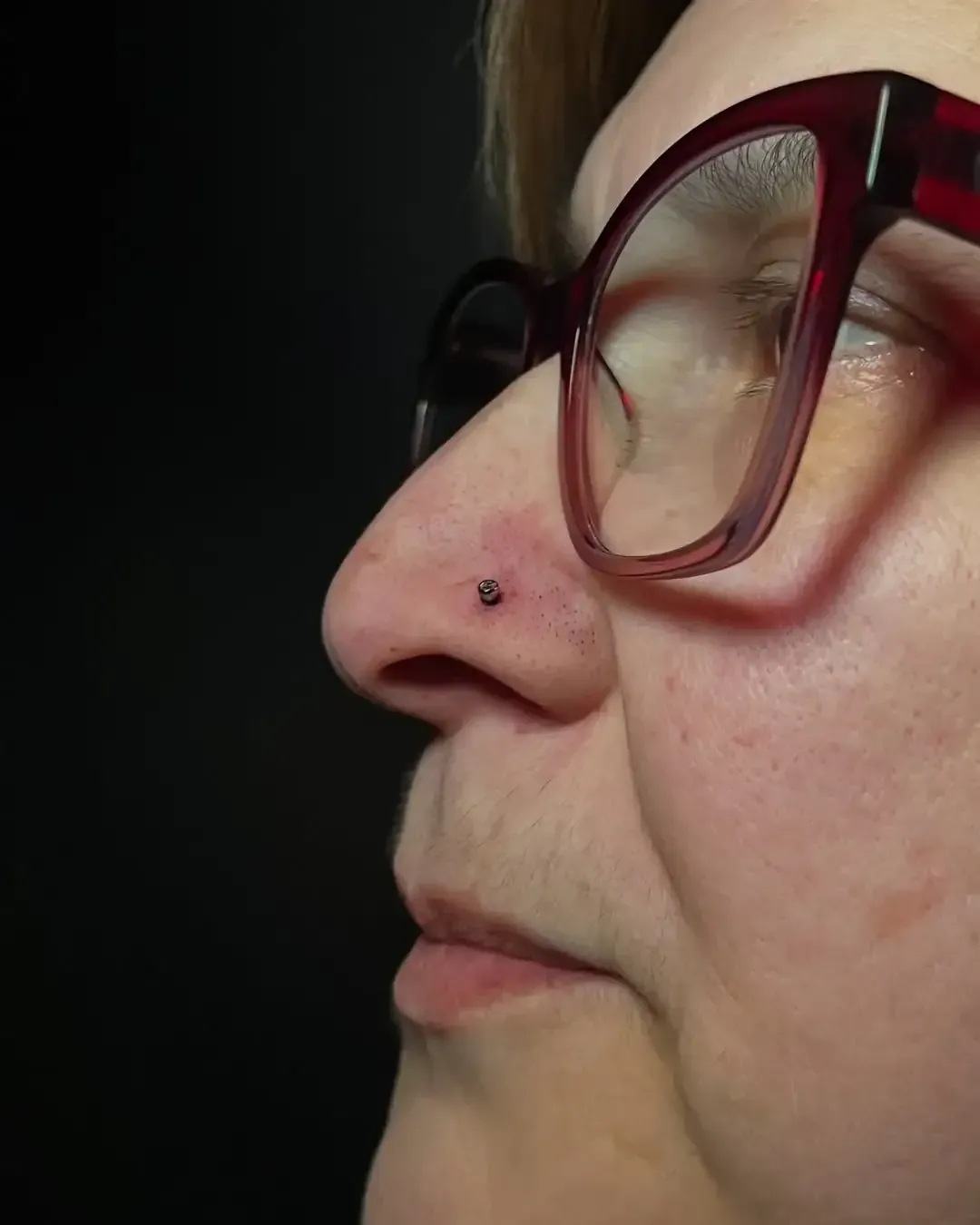 Mujer que lleva gafas con un piercing en la nariz, sobre un fondo oscuro.
