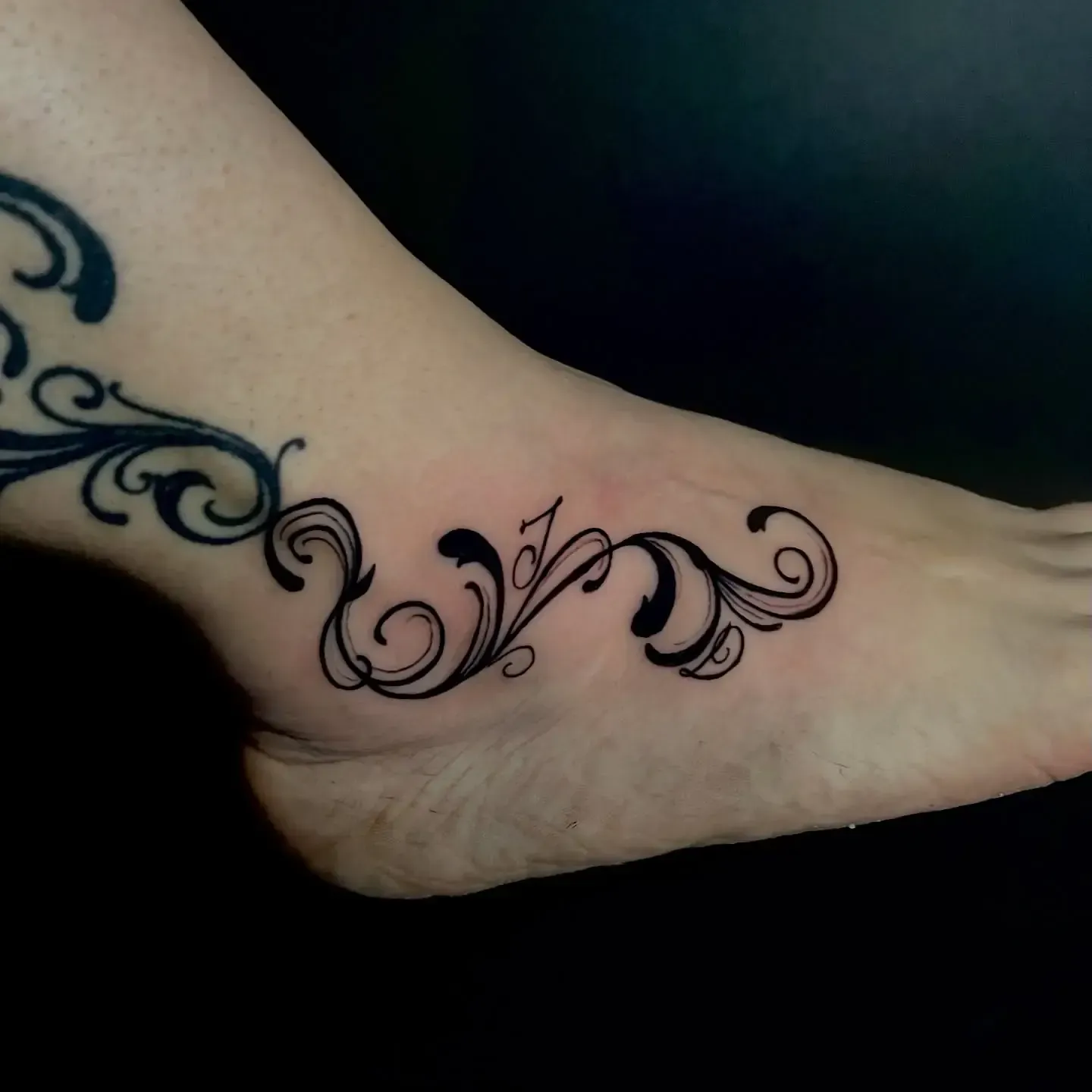 Tatuaje ornamental negro en el tobillo y el pie de una persona, sobre un fondo oscuro.