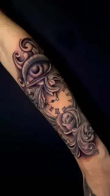 Tatuaje en el antebrazo, que representa un ojo, una esfera de reloj, una rosa y volutas ornamentadas en negro y gris.