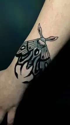 Tatuaje de polilla negra y gris en la muñeca. Alas con diseño tribal.