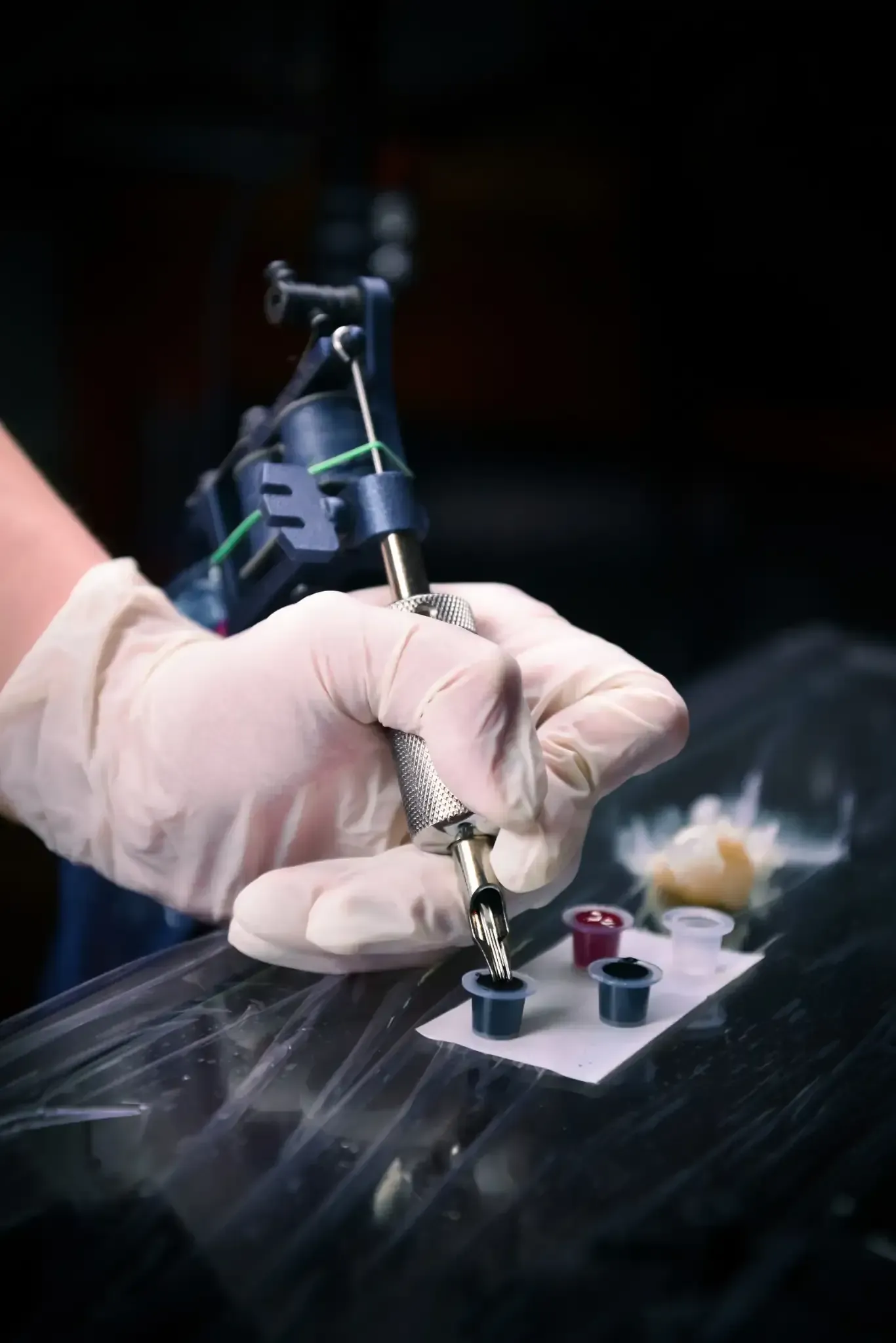 La mano enguantada de un artista del tatuaje sostiene una máquina de tatuar sobre recipientes de tinta.