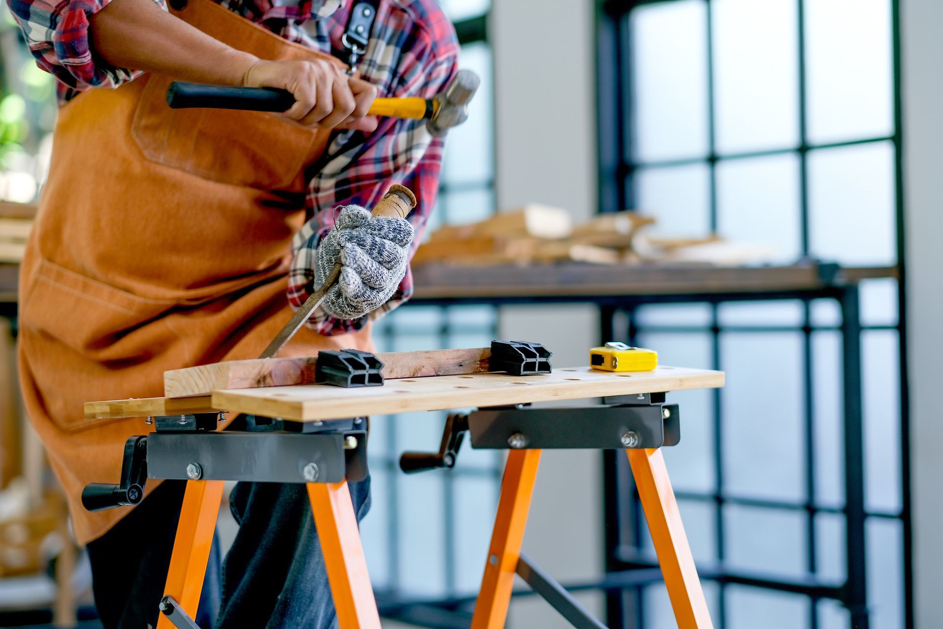 Persona martillando madera en un caballete en el taller; delantal naranja, vigas de madera, abrazaderas, ventana al fondo.