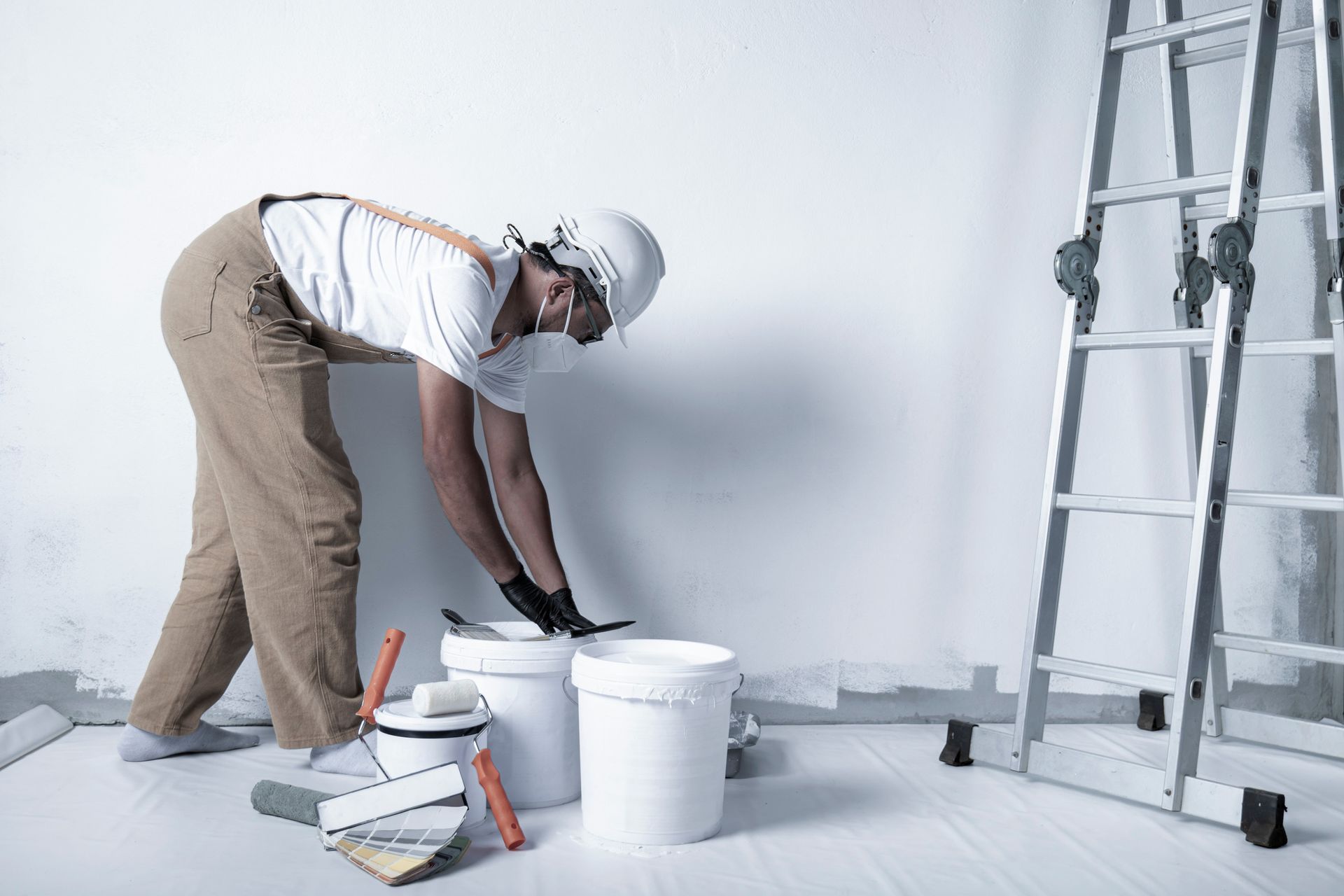 Pintor inclinado para aplicar pintura a la pared; baldes, rodillo y escalera visibles.
