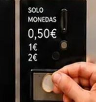 Una mano introduce una moneda en una máquina negra que funciona con monedas, con las etiquetas en español «SOLO MONEDAS» y «0,50€ 1€ 2€».