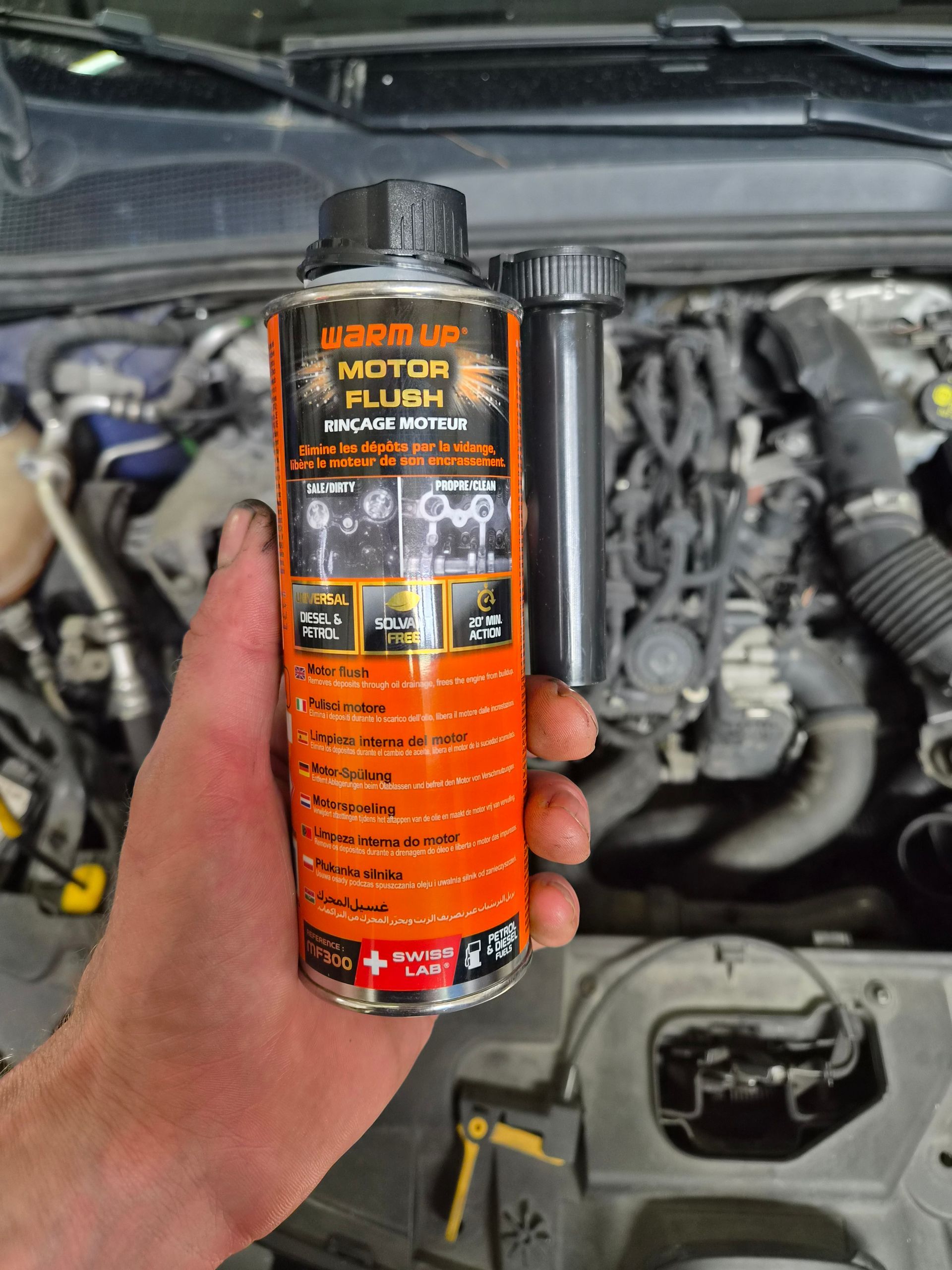 Une main tenant un flacon de traitement moteur orange dans le compartiment moteur ouvert d’une voiture