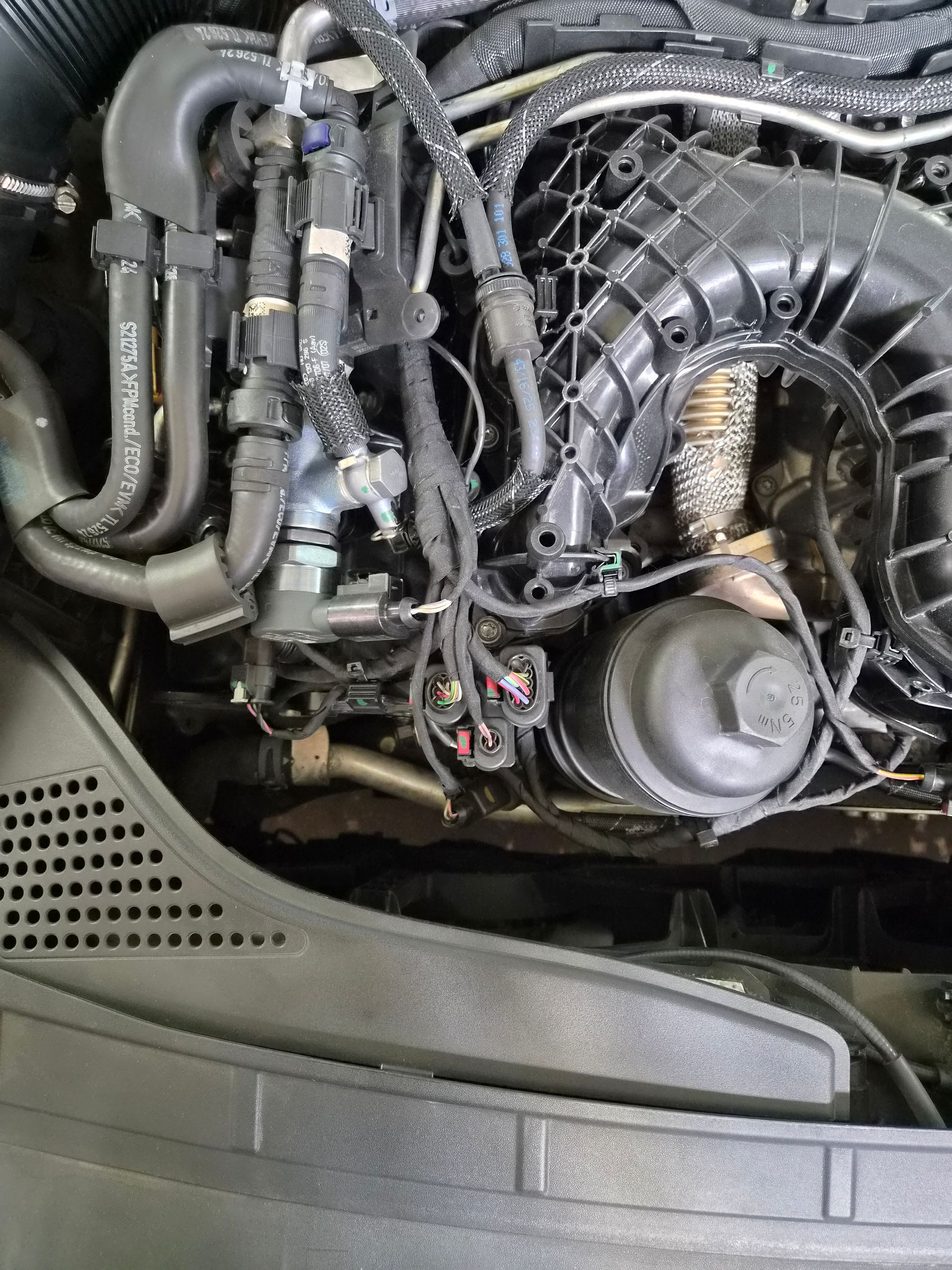 Gros plan sur le compartiment moteur d’une voiture, avec des durites noires, des câbles et un système d’admission d’air bien visible.