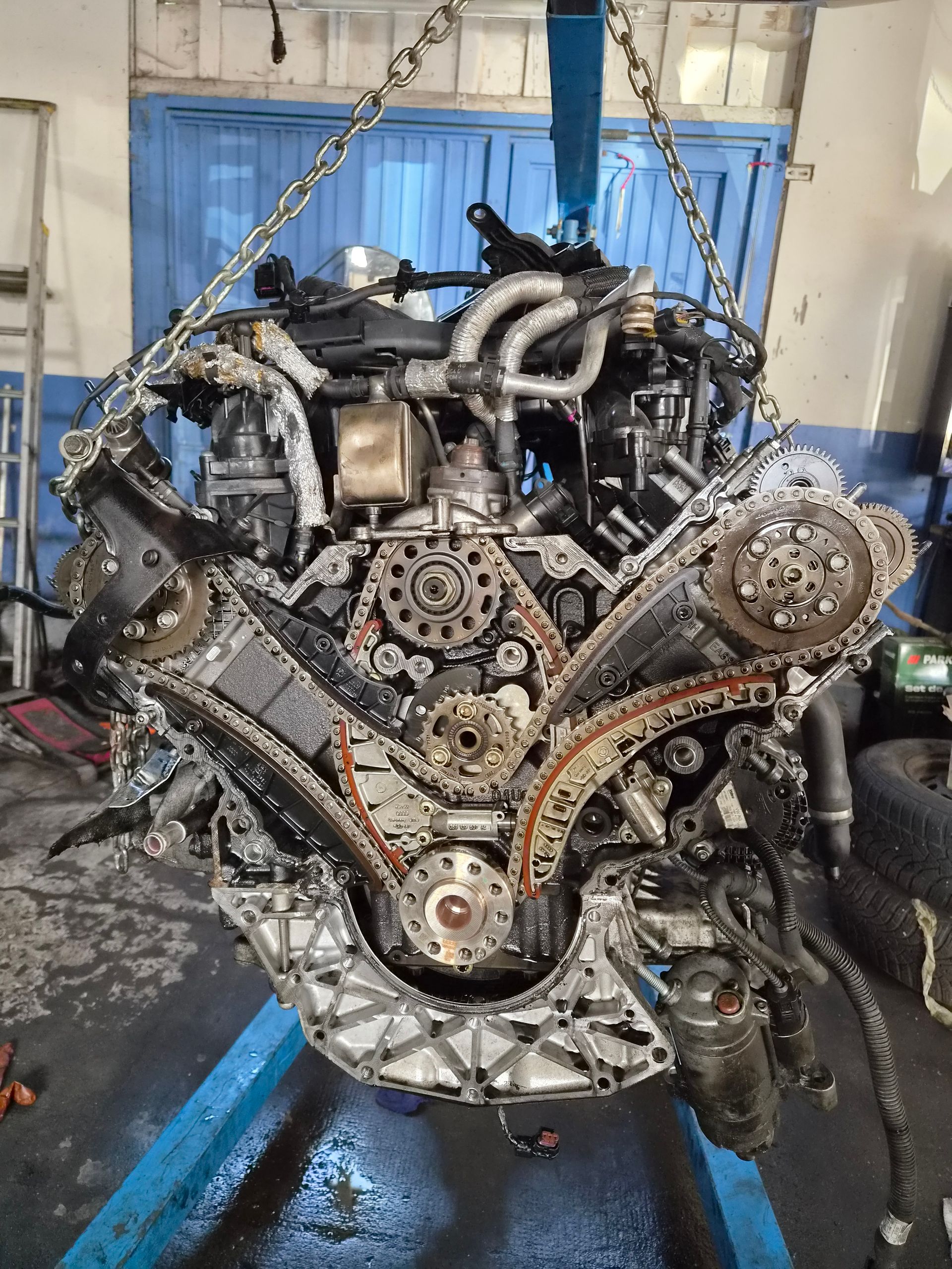 Moteur de voiture suspendu dans un atelier, avec les chaînes de distribution et les composants apparents, et le sol mouillé en dessous