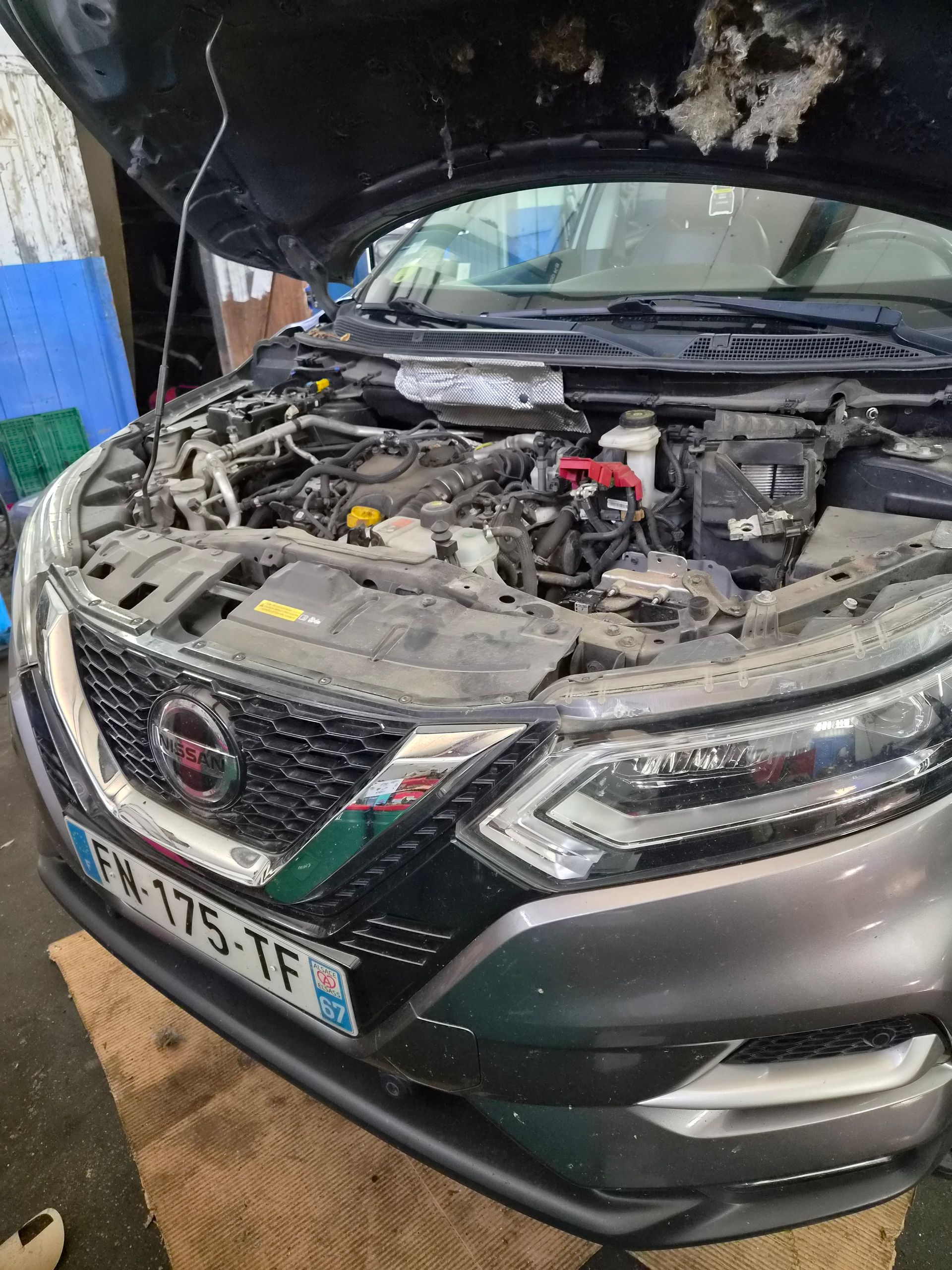 Nissan noire endommagée, capot ouvert et moteur apparent, dans un atelier avec des débris au-dessus.