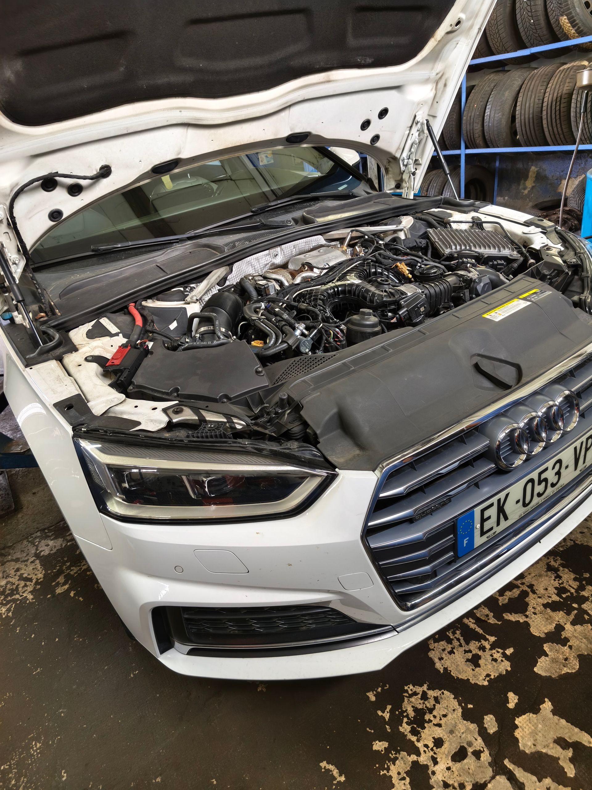 Audi blanche au capot ouvert après un choc à l’avant, à l’intérieur d’un garage, laissant apparaître le moteur et la carrosserie déformée
