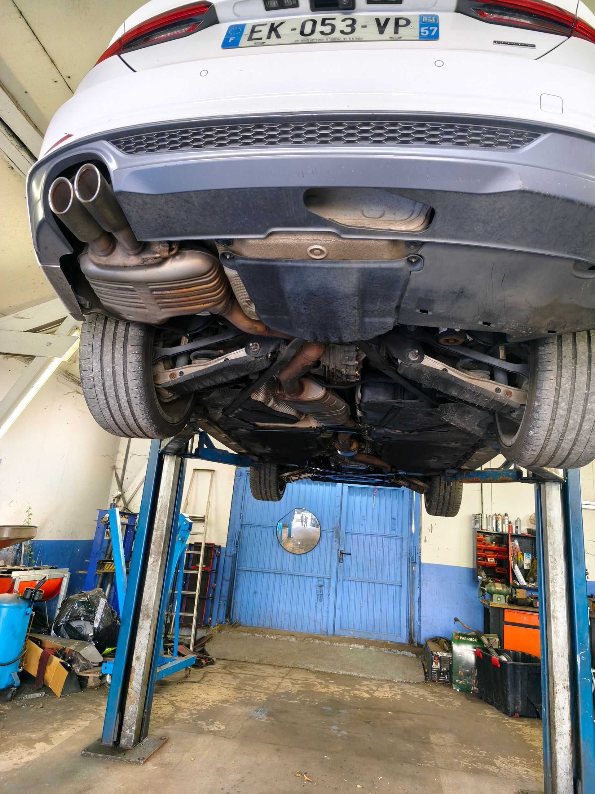 Vue du dessous d’une voiture argentée sur un pont élévateur de garage, montrant l’échappement et le châssis.