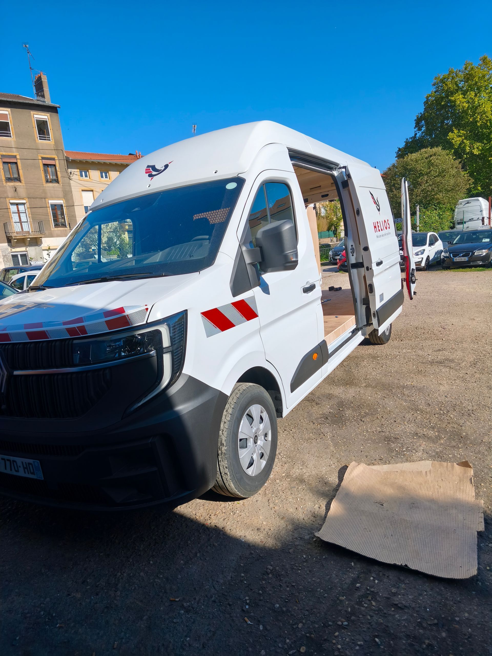 Fourgon utilitaire blanc avec la porte latérale ouverte, garé dans une rue ensoleillée près d’un bâtiment et d’arbres