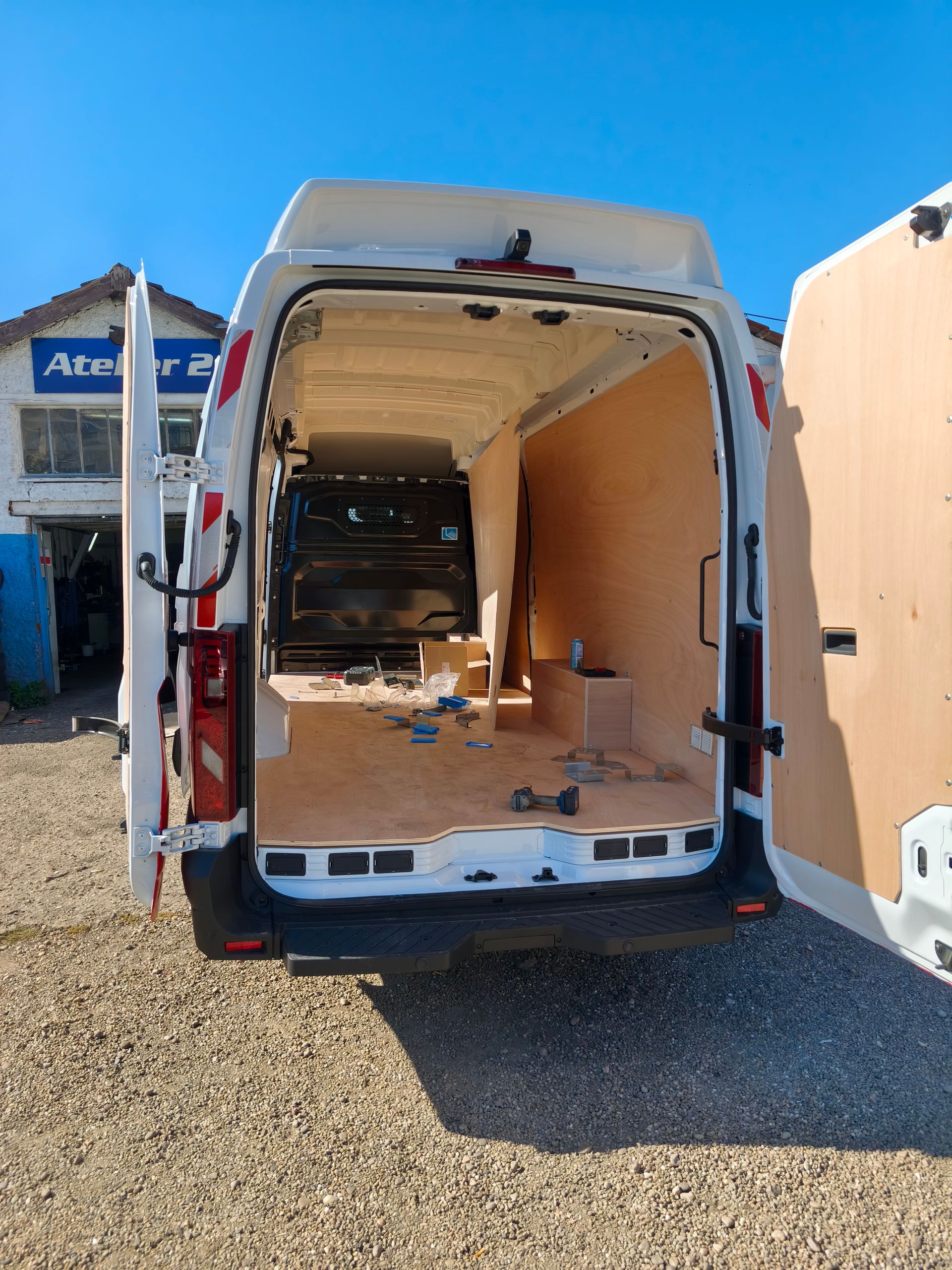 Fourgon utilitaire blanc ouvert, à l’intérieur dépouillé, stationné sur du gravier à côté de bâtiments sous un ciel bleu