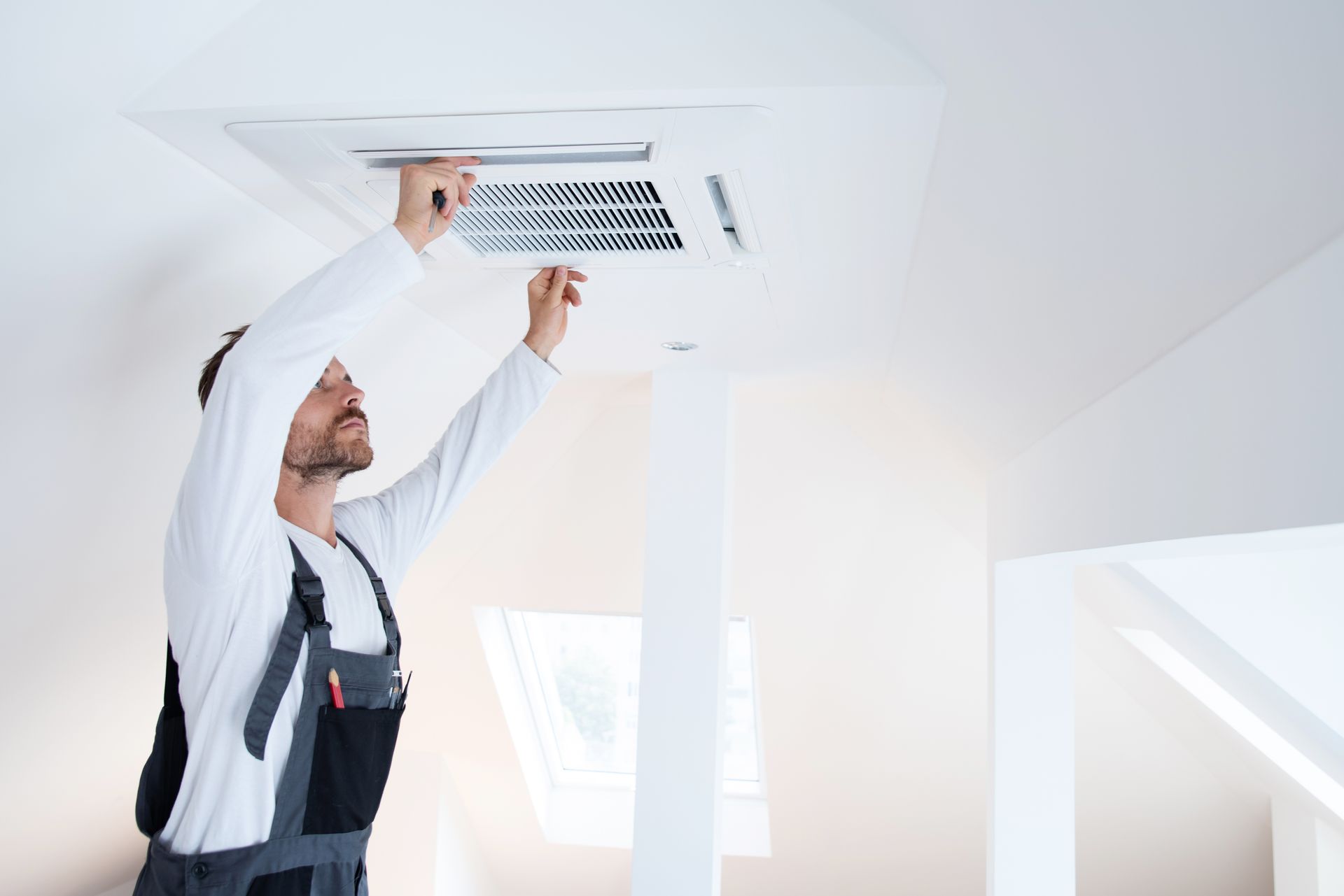 Un technicien ajuste une grille d'aération au plafond d'une pièce mansardée d'un blanc éclatant.