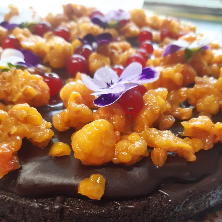 Suklaakakku, jonka päällä on karamellisoituja appelsiiniryppäitä, marjoja ja violetti syötävä kukka