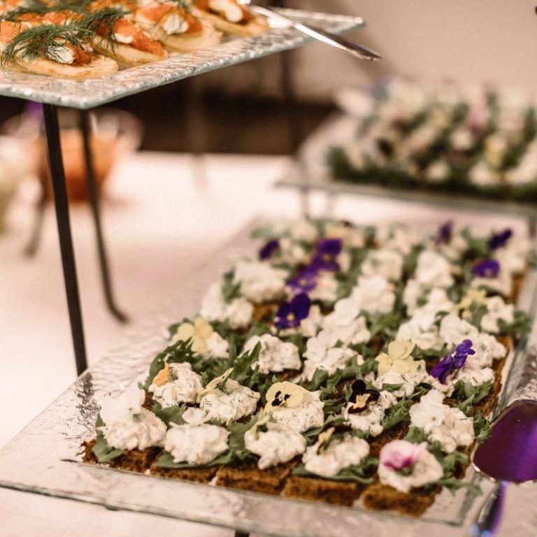 Pöydällä noutopöytätarjottimilla pieniä alkupaloja, joiden päällä on valkoista kermaa ja violetti koriste