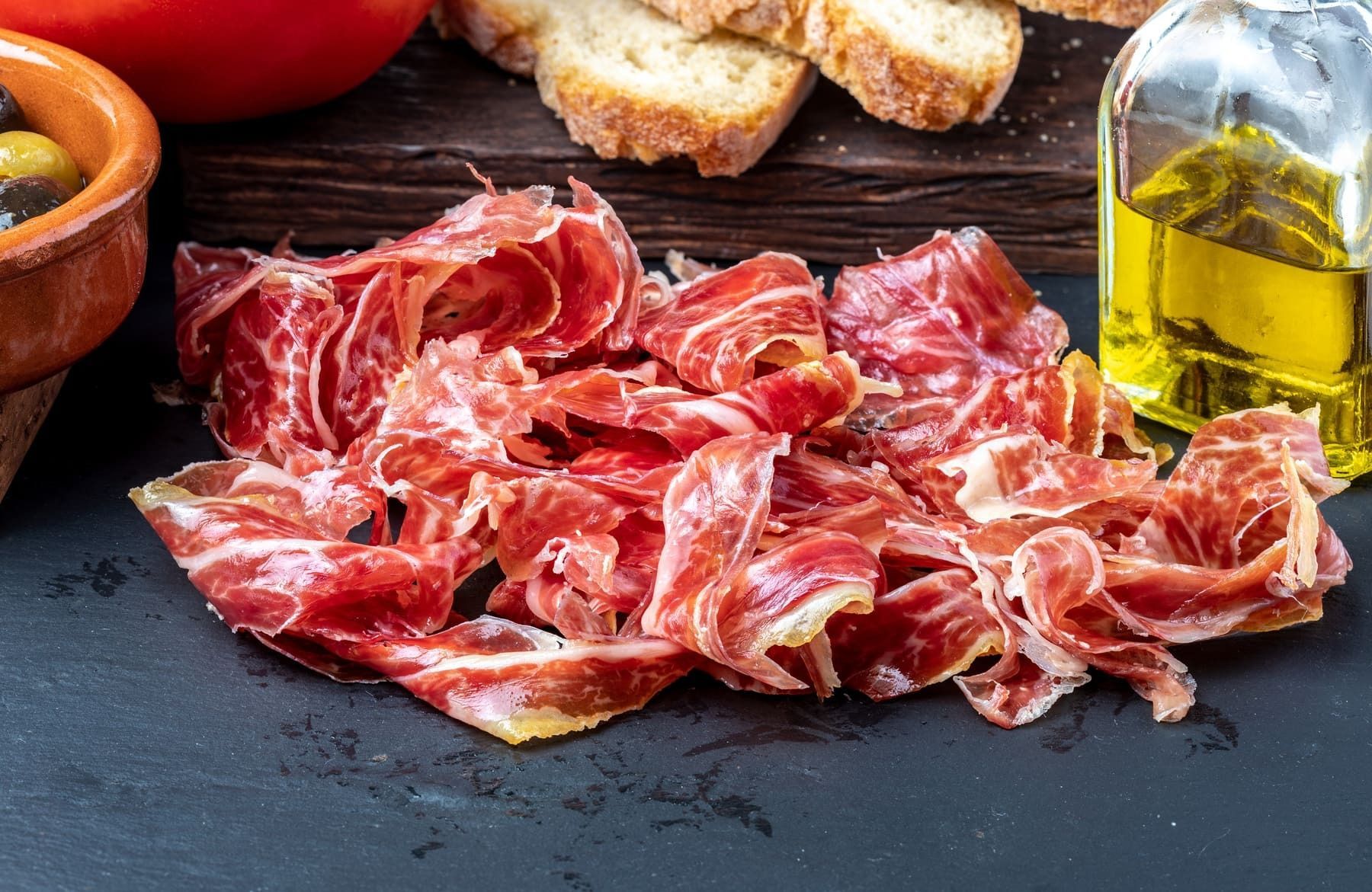 Una tabla de cortar cubierta con jamón en rodajas, aceitunas, tomates y una botella de aceite de oliva.
