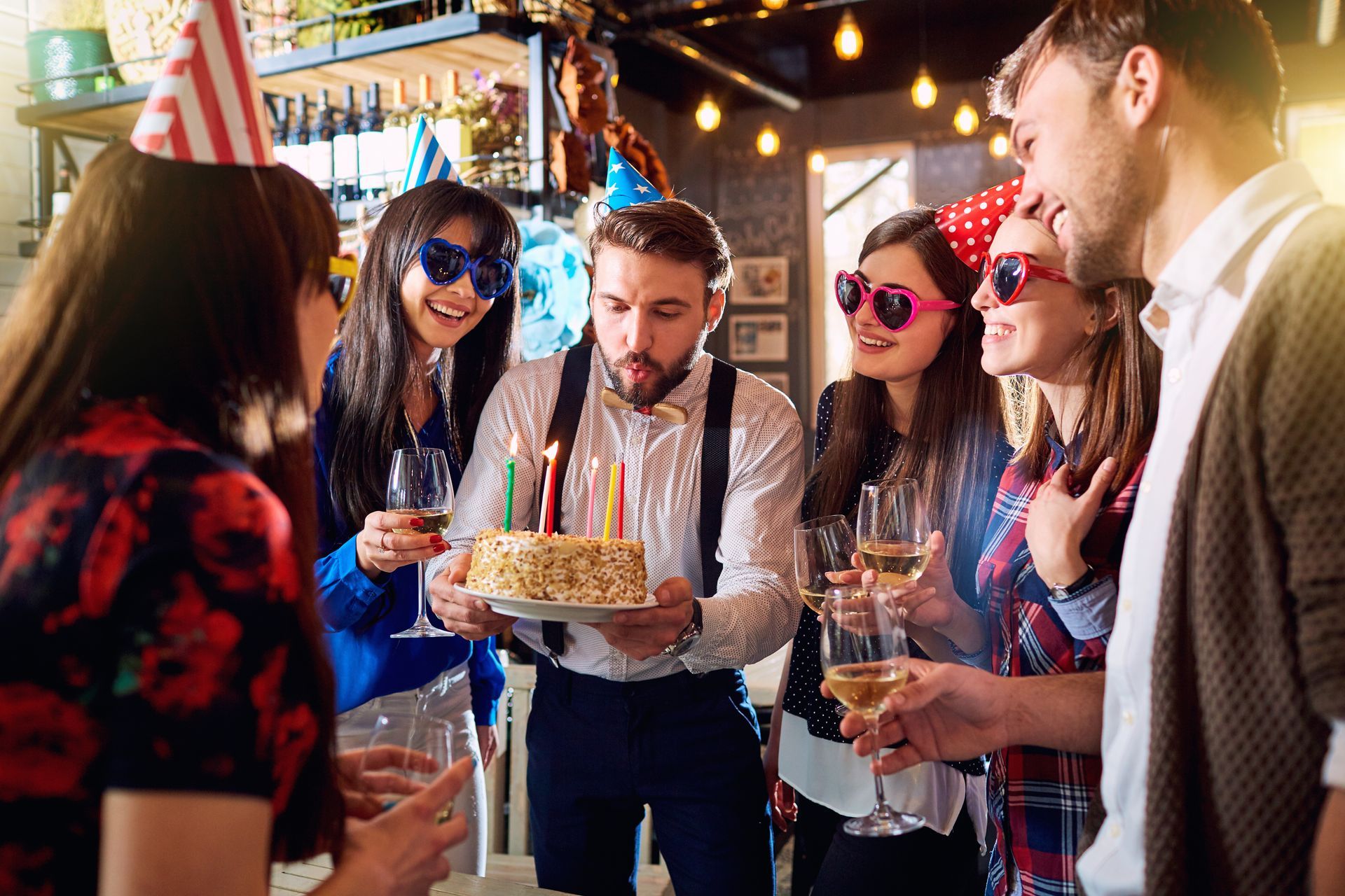 Des amis se sont réunis autour d'un gâteau d'anniversaire lors d'une fête en intérieur, avec des guirlandes lumineuses et des boissons.