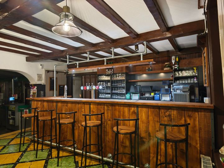 Intérieur d'un bar chaleureux avec tabourets en bois, poutres apparentes et bouteilles derrière le comptoir.