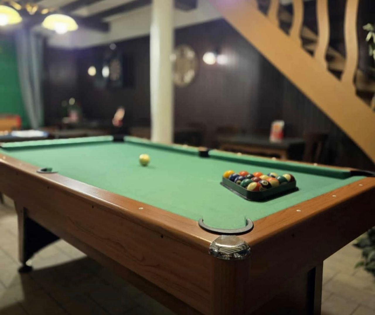 Table de billard et boules éparpillées dans une pièce faiblement éclairée, un escalier en arrière-plan.