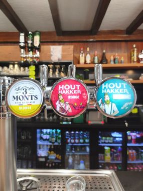 Trois tireuses à bière Houtkaker dans un bar avec des bouteilles et des réfrigérateurs derrière elles.
