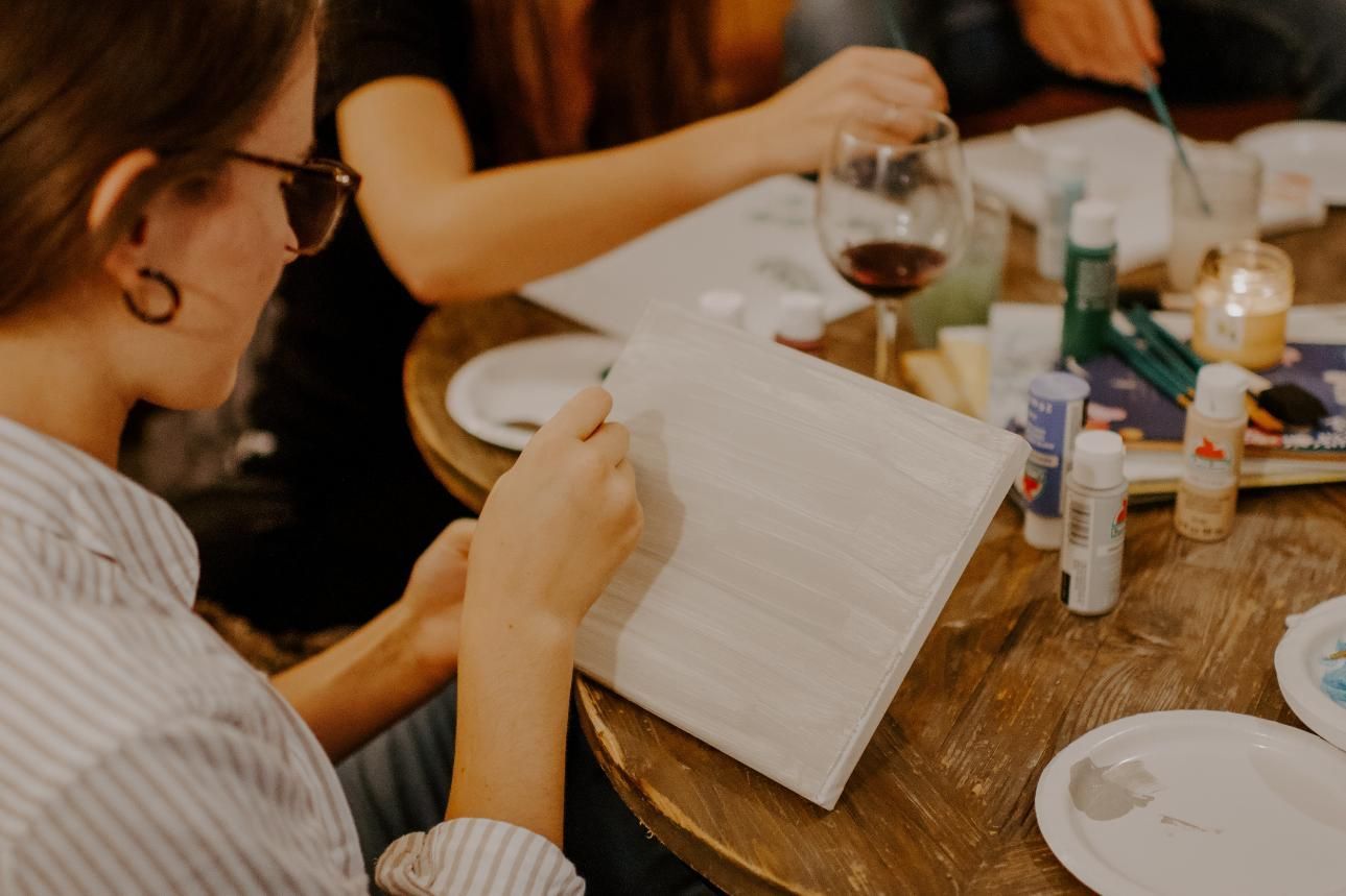 Gente pintando sobre lienzos en una mesa, con suministros de pintura y vino.
