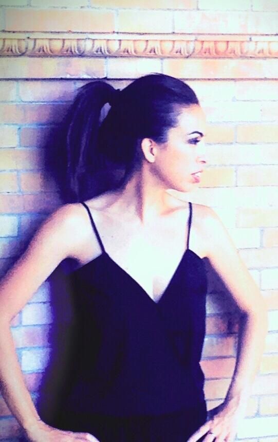 Mujer con cola de caballo en vestido negro apoyada contra una pared de ladrillos.