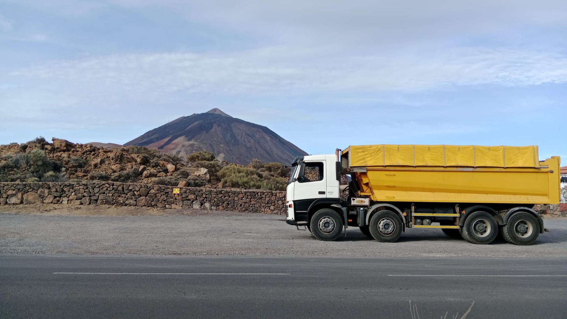 Camión volquete amarillo estacionado en una carretera junto a un terreno rocoso con una montaña al fondo.