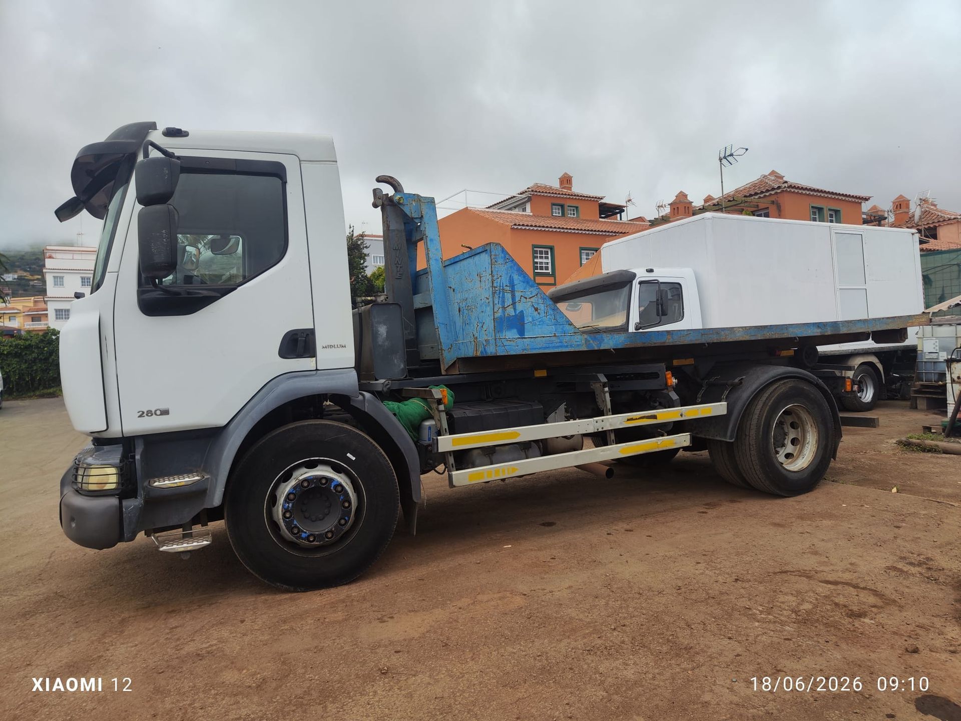 Camión blanco de plataforma con brazo de grúa azul y caja de carga blanca estacionado en un terreno de tierra.