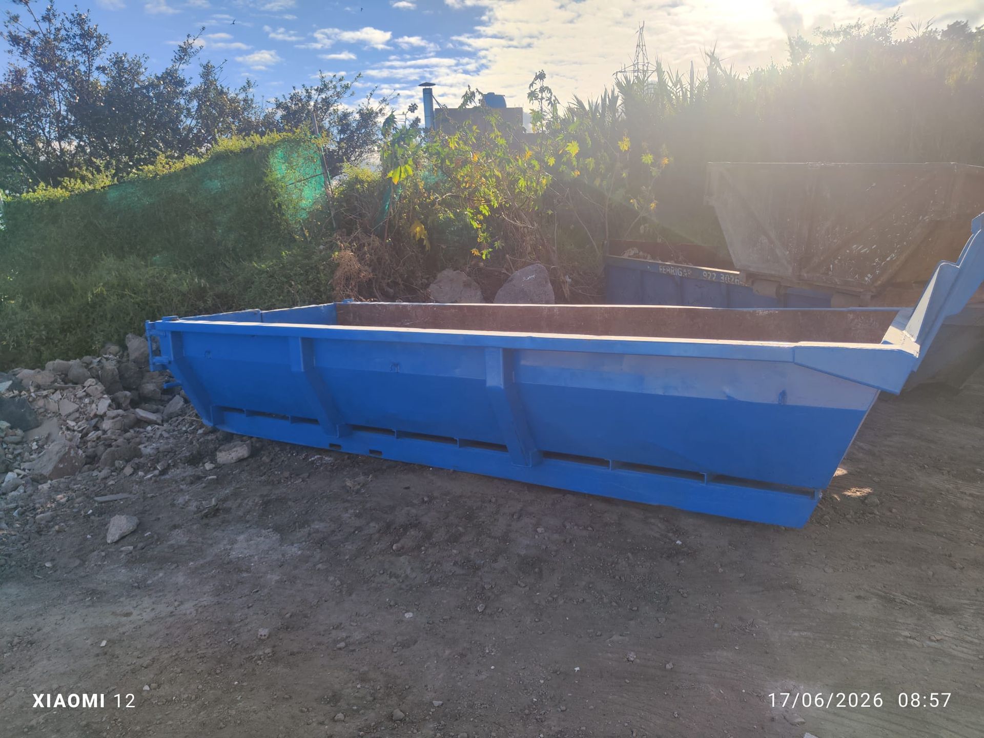 Un contenedor de basura metálico azul sobre la tierra junto a unos arbustos, bajo la luz brillante del sol
