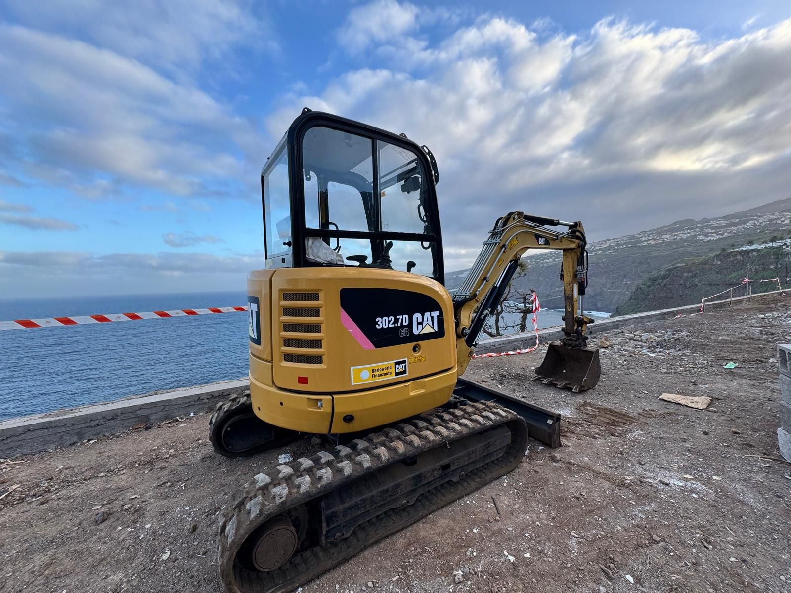 Excavadora amarilla en terreno rocoso costero junto al mar bajo un cielo nublado.
