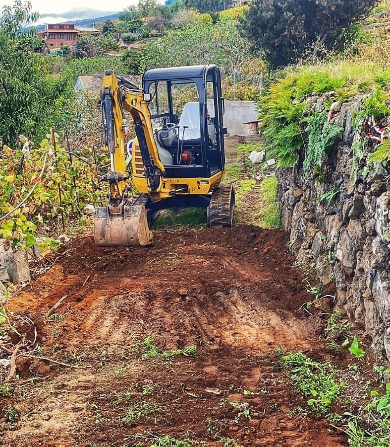 Una excavadora amarilla despeja un camino de tierra junto a un terraplén rocoso en una zona rural verde.