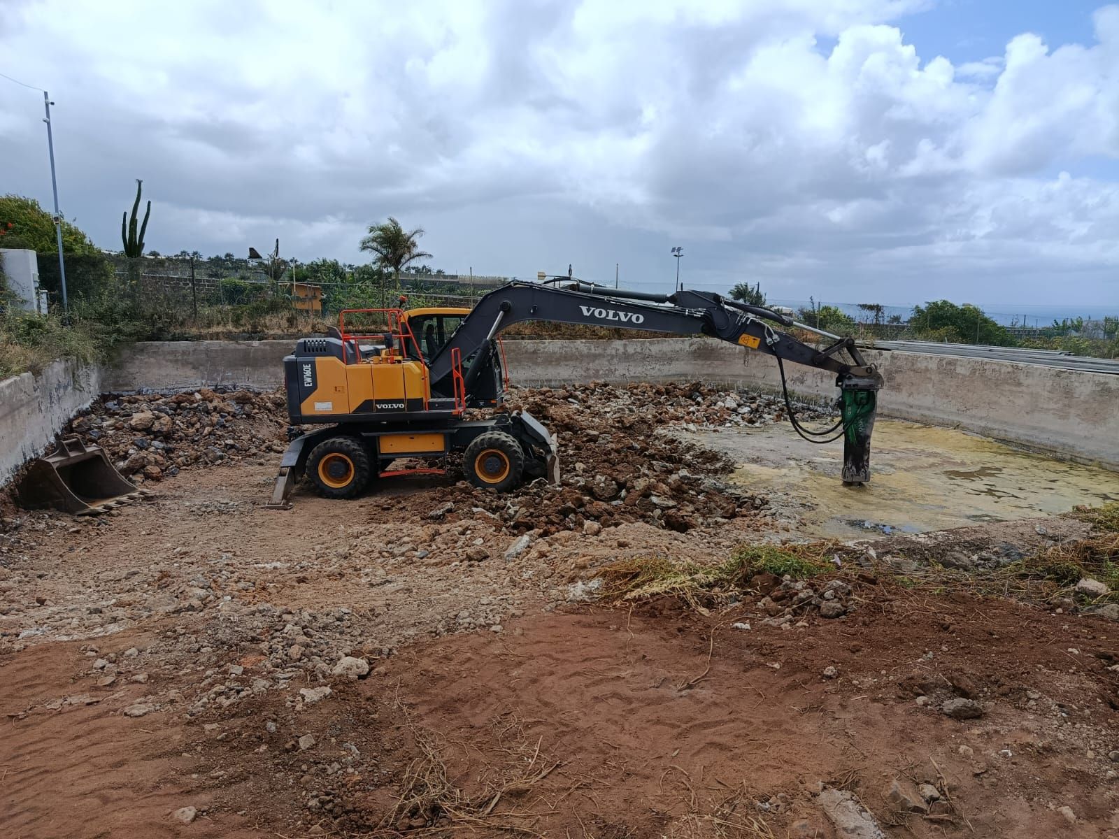 Una excavadora amarilla retira tierra en una obra fangosa bajo un cielo nublado.