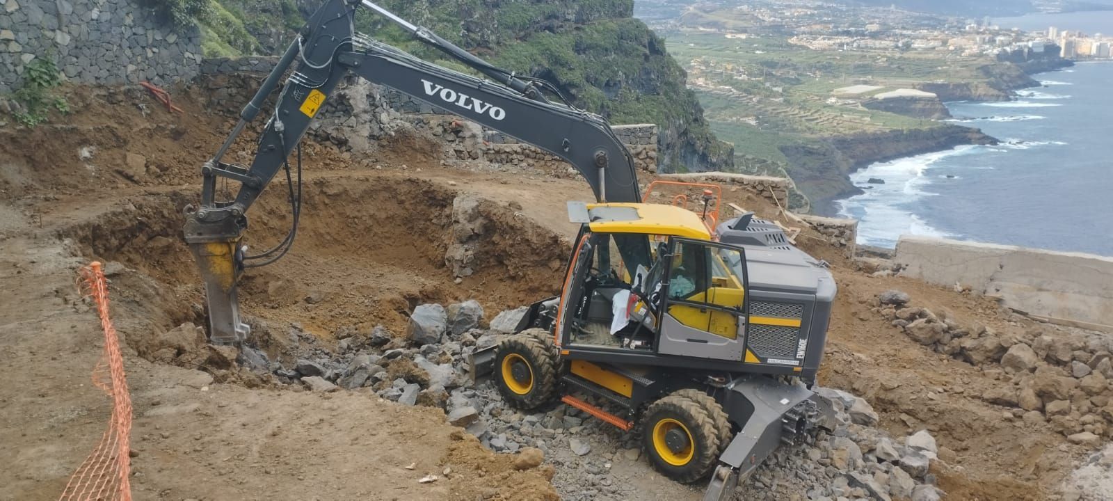 Excavadora amarilla removiendo tierra en un acantilado rocoso, con el océano al fondo.