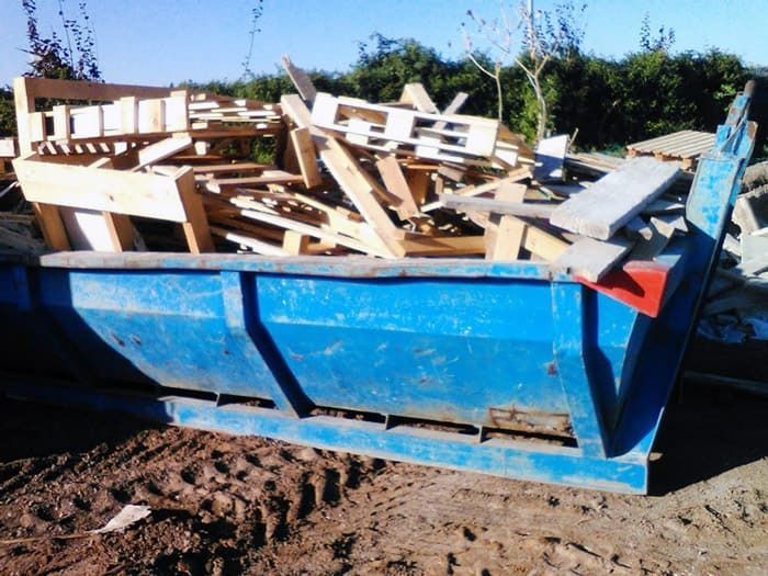 Un contenedor de basura azul lleno de madera está tirado en la tierra.