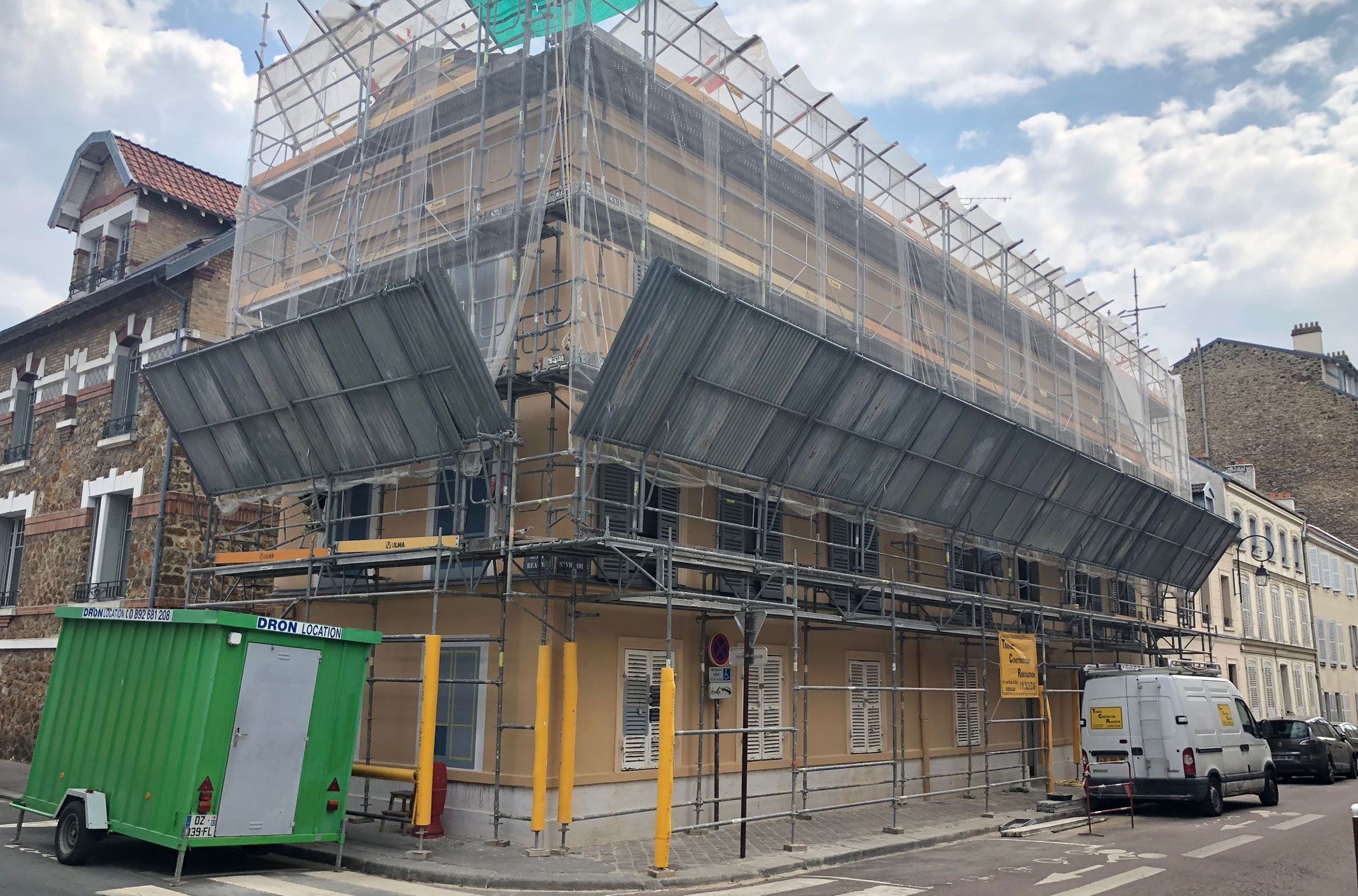 Échafaudages et filets autour d'un immeuble en rénovation, avec une benne à déchets verte dans la rue.