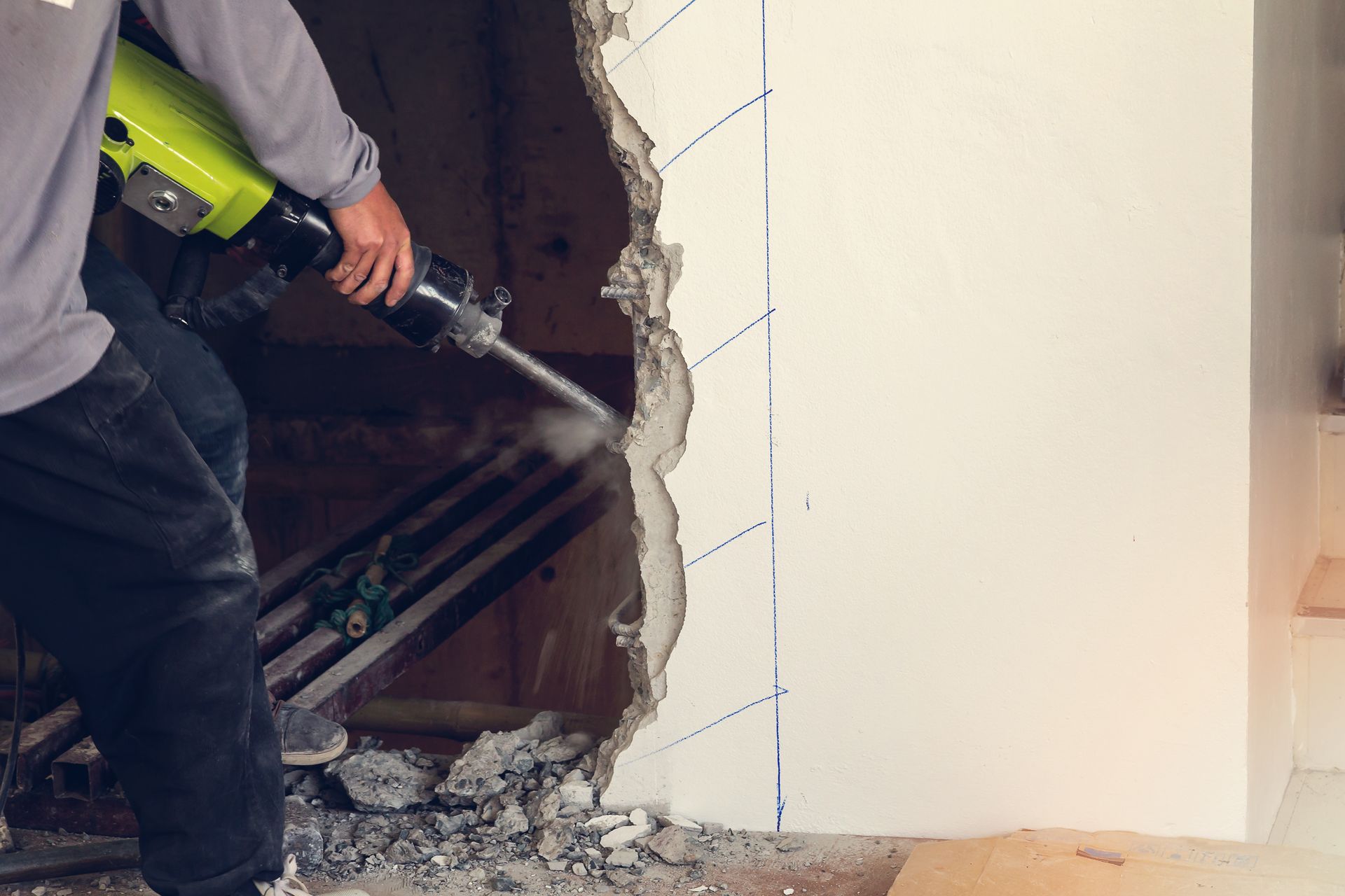 Un ouvrier utilise un marteau-piqueur pour démolir un mur ; des gravats et de la poussière jonchent le sol.