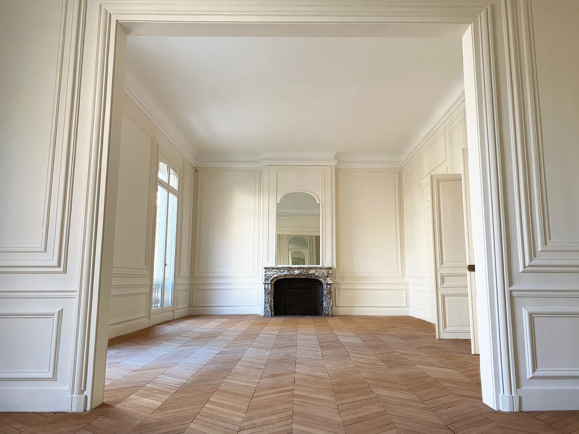 Élégante pièce, ornée de moulures, d'un parquet à chevrons et d'une cheminée noire centrée sur le mur du fond.