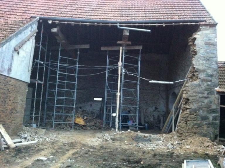 Bâtiment en pierre partiellement démoli, façade ouverte, échafaudages et débris sous un toit de tuiles