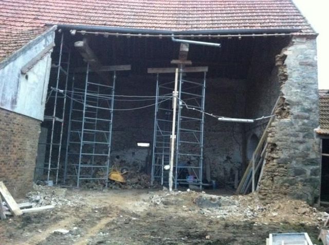 Bâtiment en pierre partiellement démoli, façade ouverte, échafaudages et débris sous un toit de tuiles