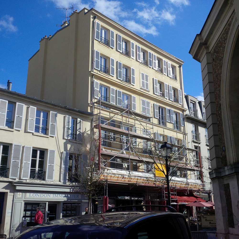 Rue avec un grand immeuble beige, des devantures de magasins et des piétons sous des échafaudages