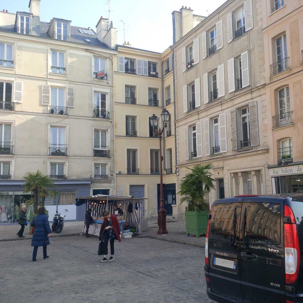 Place parisienne avec des immeubles beiges, des piétons et des véhicules garés.