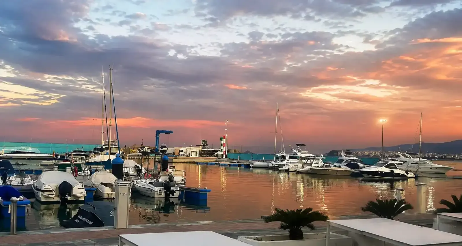 Puesta de sol sobre un puerto deportivo con yates, barcos y luces del puerto reflejadas en aguas tranquilas.