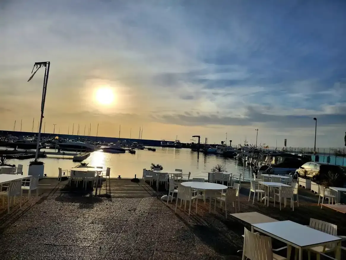 Puesta de sol sobre un puerto deportivo con barcos atracados, aguas tranquilas y mesas de café a lo largo del paseo marítimo.