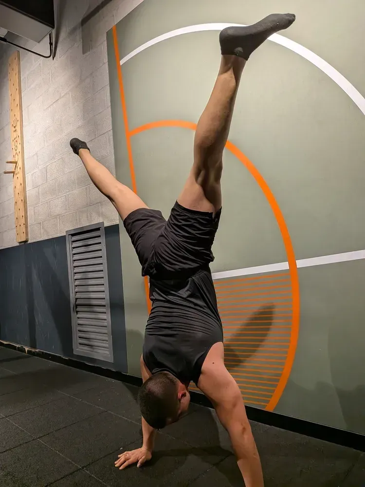Persona haciendo parada de manos apoyada contra una pared, con una pierna levantada, vistiendo pantalones cortos negros y camiseta sin mangas.