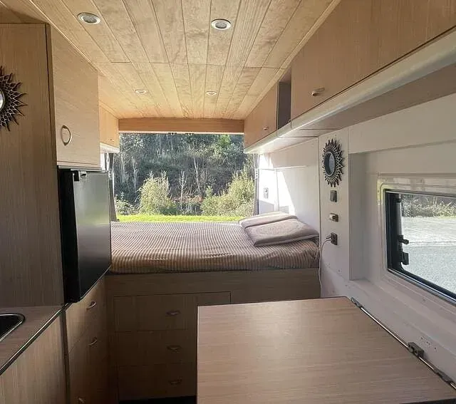 Vista interior de una autocaravana con cama, cocina y mesa. La autocaravana tiene paneles de madera clara y vistas a un entorno natural.