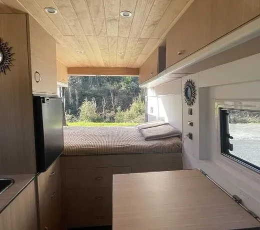 Vista interior de una autocaravana con cama, cocina y mesa. La autocaravana tiene paneles de madera clara y vistas a un entorno natural.